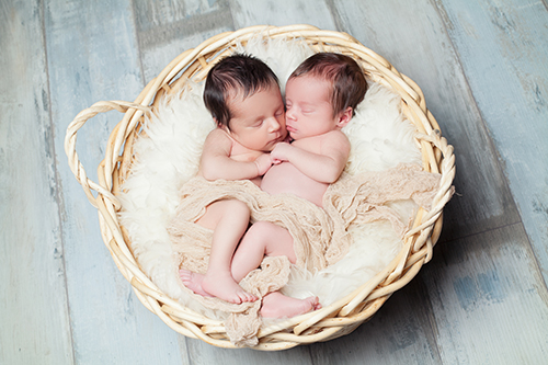 Fotografie Neugeborene Düsseldorf: Baby-Zwillinge liegen in weißer Decke aneinandergekuschelt in Körbchen und schlafen.