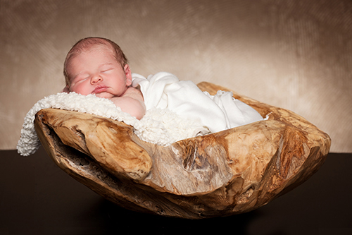 Neugeborenenfotos Düsseldorf: Baby in weißer Wolldecke liegt in einer dicken Holzschale vor braunem Hintergrund.