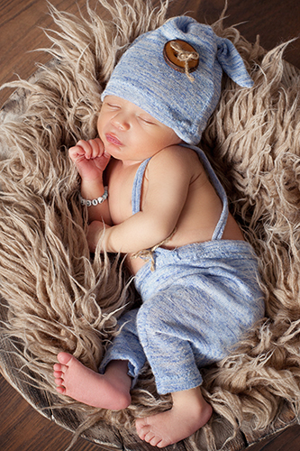 Babyfotos Düsseldorf: Baby mit blauer Hose und blauer Mütze liegt seitlich auf brauner Zottel-Wolldecke