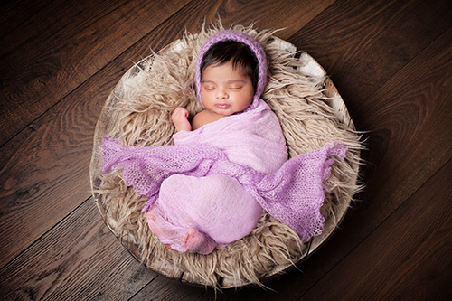 Fotostudio Baby Duesseldorf: Baby mit pechschwarzem Haar schkummert in lila Tuch gewickelt in einer mit Wolldecke ausgelegten Holzschale